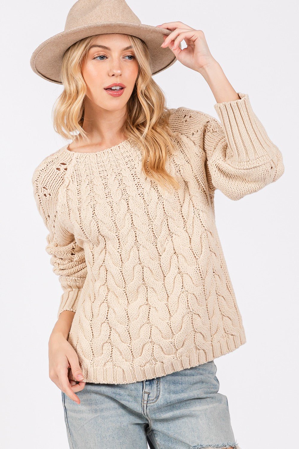 A cozy cable knit long sleeve sweater in a soft cream color featuring a textured pattern and ribbed cuffs displayed on a wooden hanger against a neutral background.