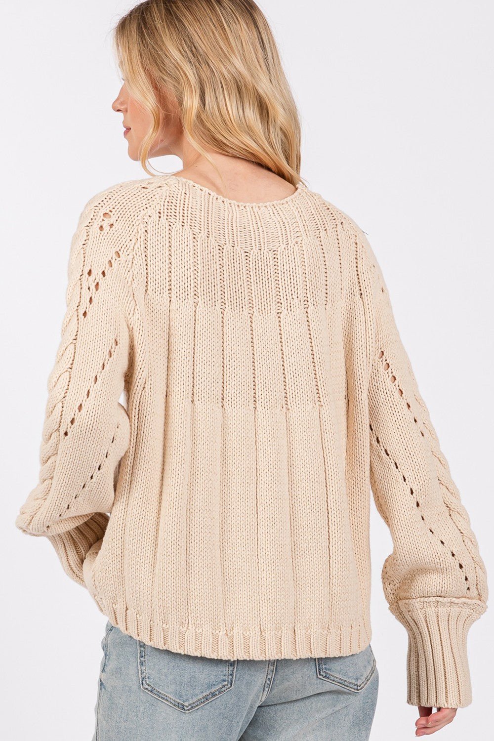 A cozy cable knit long sleeve sweater in a soft cream color featuring a textured pattern and ribbed cuffs displayed on a wooden hanger against a neutral background.